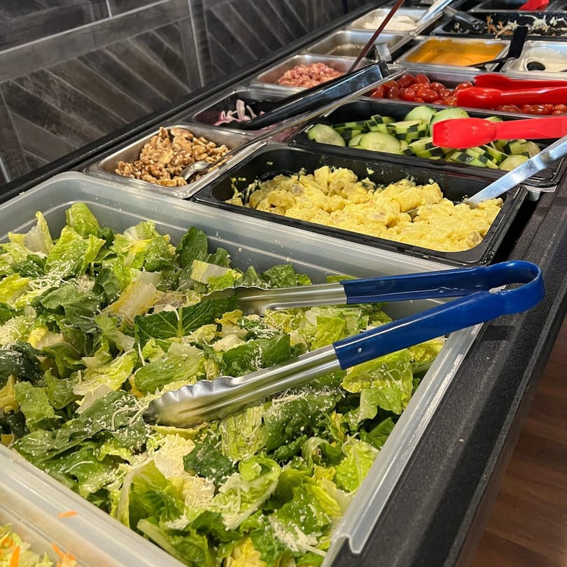 A salad bar with Caesar salad, potato salad, walnuts, cucumbers, cherry tomatoes, and other toppings.