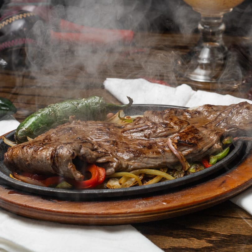 Sizzling Mi Casita fajitas with steak, onions, and peppers, emitting steam on a cast iron skillet.