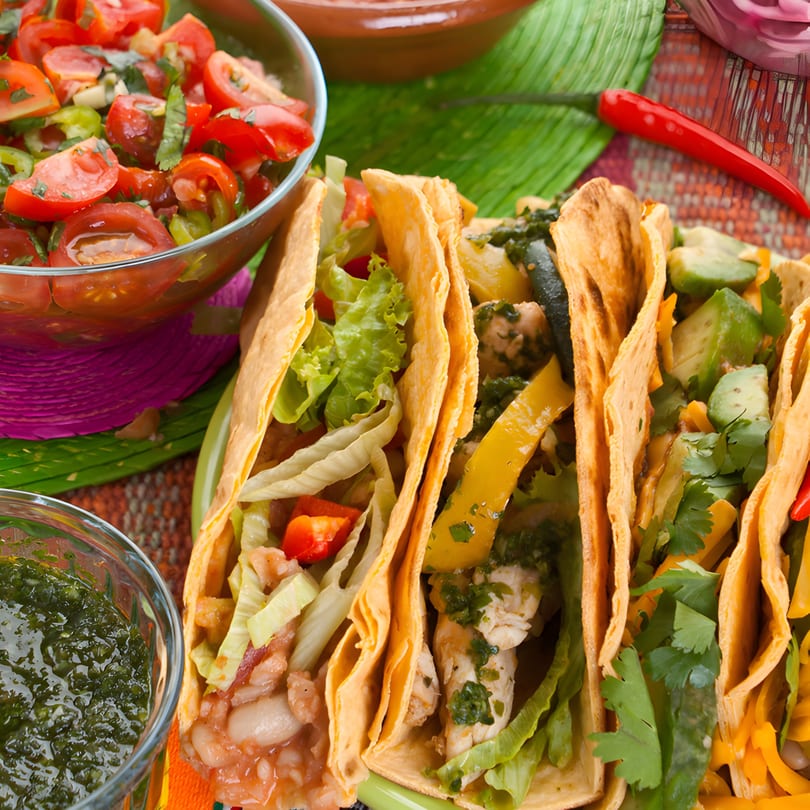 Three Mi Casita tacos with various fillings, salsa, and green sauce on a colorful Mexican-themed table.