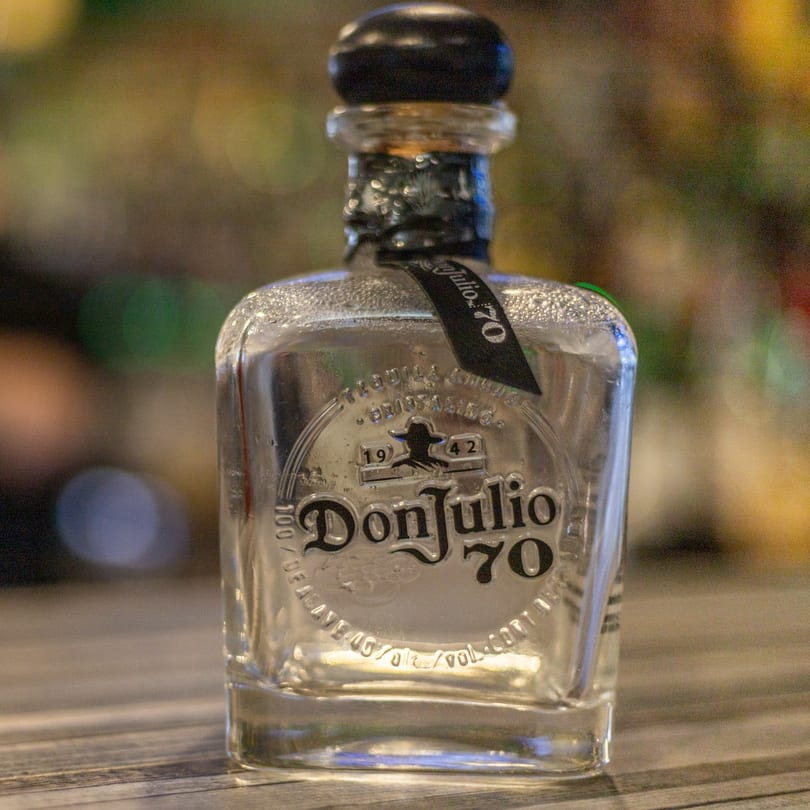 A bottle of Don Julio 70 Tequila, clear with a black label, sits on a wooden surface.